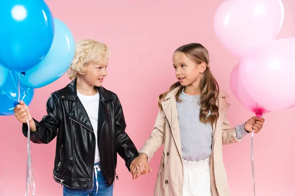 Niños sonrientes sosteniendo globos y tomándose de las manos sobre fondo rosa - foto de stock