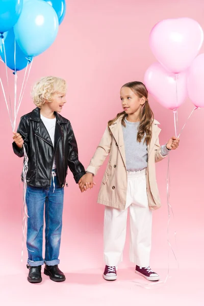 Niños sonrientes sosteniendo globos y tomándose de las manos sobre fondo rosa - foto de stock