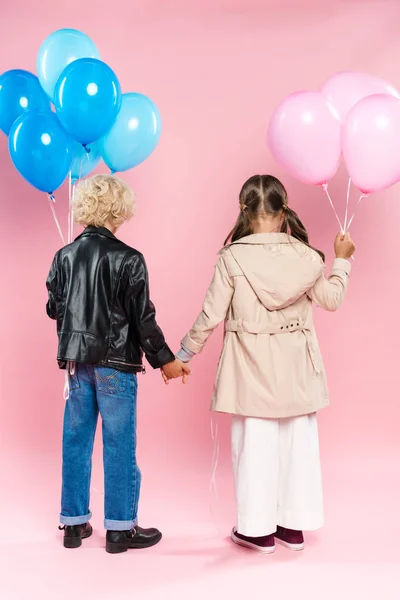 Vista posterior de los niños sosteniendo globos y tomándose de las manos sobre fondo rosa - foto de stock