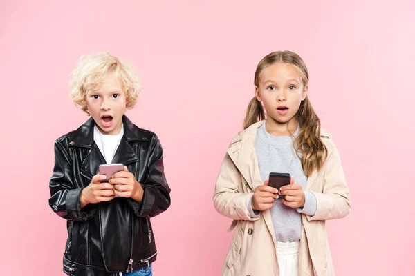 Crianças chocadas segurando smartphones e olhando para a câmera no fundo rosa — Fotografia de Stock