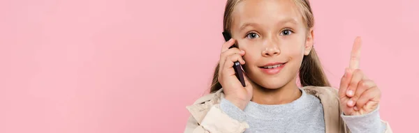 Plano panorámico de niño sonriente mostrando signo de idea y hablando en el teléfono inteligente aislado en rosa - foto de stock