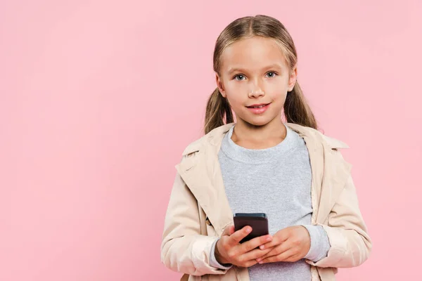 Niño sonriente en traje de otoño sosteniendo teléfono inteligente aislado en rosa - foto de stock