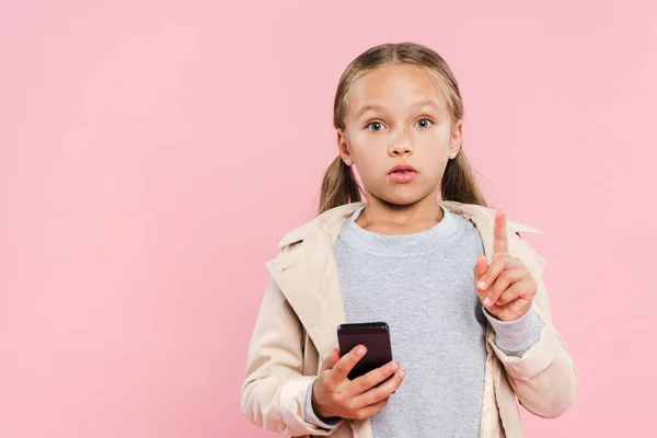 Шокована дитина показує знак ідеї і тримає смартфон ізольований на рожевому — стокове фото