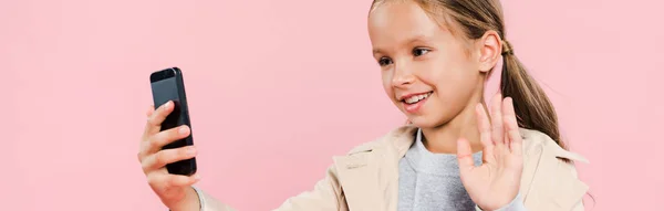 Панорамный снимок улыбающегося и милого ребенка, машущего во время видеочата, изолированного на розовом — стоковое фото