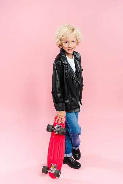 Sonriente y lindo niño sosteniendo penny board sobre fondo rosa - foto de stock