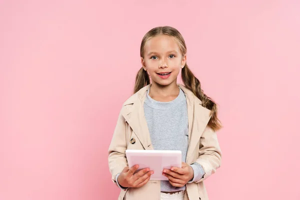 Niño sonriente sosteniendo la tableta digital y mirando a la cámara aislada en rosa - foto de stock