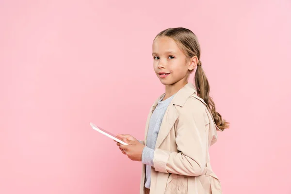 Усміхнена дитина тримає цифровий планшет і дивиться на камеру ізольовано на рожевому — стокове фото