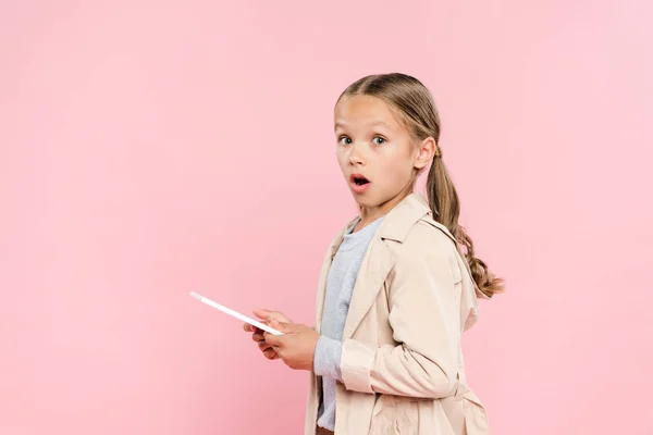 Schockiertes Kind hält digitales Tablet in der Hand und blickt isoliert auf Kamera in rosa — Stockfoto