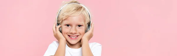 Panoramaaufnahme eines lächelnden Kindes mit Kopfhörern, das isoliert auf rosa Musik hört — Stockfoto