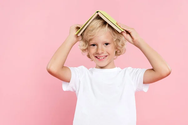Bambino sorridente che tiene il libro e guarda la fotocamera isolata sul rosa — Foto stock