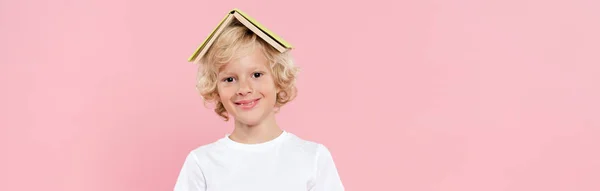 Panoramic shot of smiling kid with book on head isolated on pink — Stock Photo