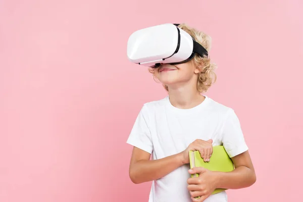 Criança sorridente com realidade virtual headset segurando livro isolado em rosa — Fotografia de Stock