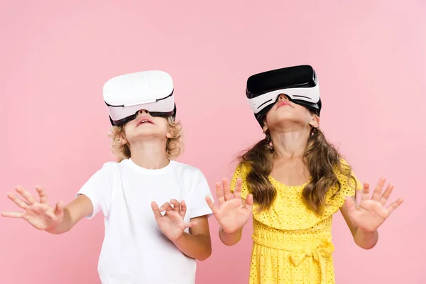 Crianças com fone de ouvido realidade virtual e mãos estendidas olhando para cima no fundo rosa — Fotografia de Stock