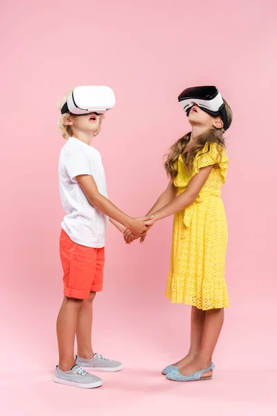 Shocked kids with virtual reality headset holding hands on pink background — Stock Photo