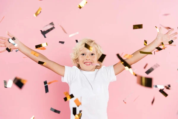 Niño sonriente con las manos extendidas cerca de caer confeti sobre fondo rosa - foto de stock