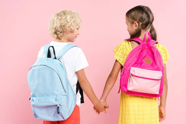 Vista posterior de los escolares con mochilas cogidas de la mano y mirándose - foto de stock