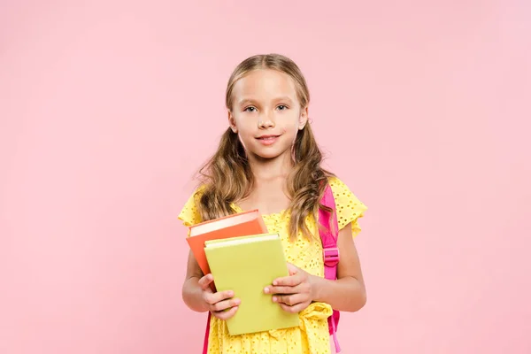 Scolaretta sorridente con zaino che tiene libri isolati su rosa — Foto stock