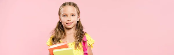 Panoramaaufnahme eines lächelnden Schulmädchens mit Rucksack, das Bücher isoliert auf rosa Papier hält — Stockfoto