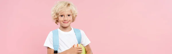 Panoramaaufnahme eines lächelnden Schuljungen mit Rucksack, der Bücher isoliert auf rosa Papier hält — Stockfoto