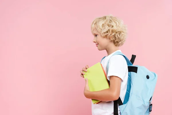 Вид збоку усміхненого школяра з рюкзаком тримає книгу ізольовано на рожевому — стокове фото