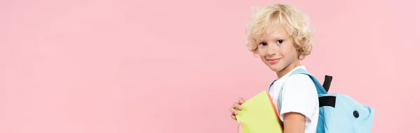 Panoramic shot of smiling schoolboy with backpack holding book isolated on pink — Stock Photo