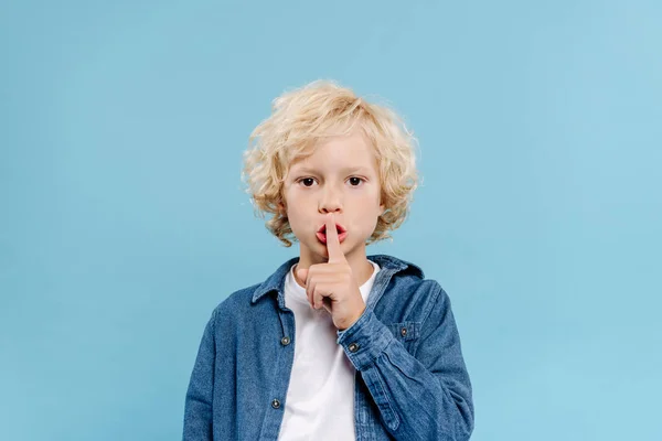 Lindo niño mostrando gesto secreto y mirando a la cámara aislada en azul - foto de stock