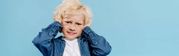 Panoramaaufnahme eines irritierten und niedlichen Kindes, das die Ohren berührt und isoliert auf blau in die Kamera blickt — Stockfoto