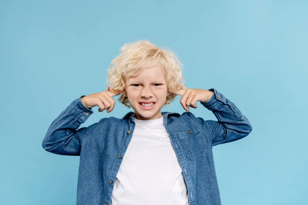 Niño irritado y lindo mirando a la cámara y tocando las orejas aisladas en azul - foto de stock