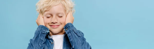 Plano panorámico de niño sonriente y lindo con los ojos cerrados tocando las orejas aisladas en azul - foto de stock