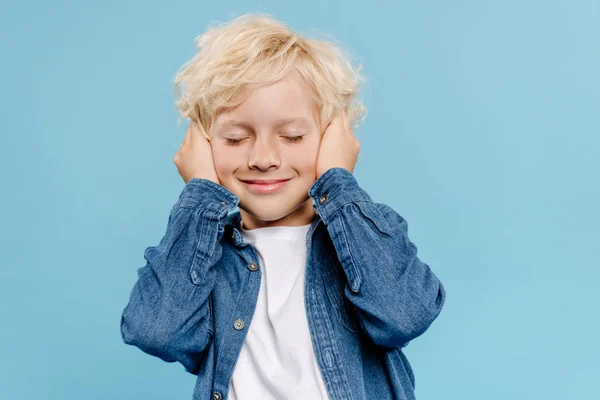 Smiling and cute kid with closed eyes touching ears isolated on blue — Stock Photo