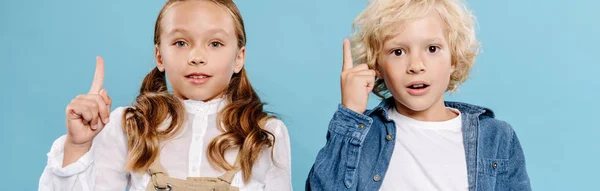 Panoramic shot of shocked and cute kids looking at camera and showing idea sign isolated on blue — Stock Photo