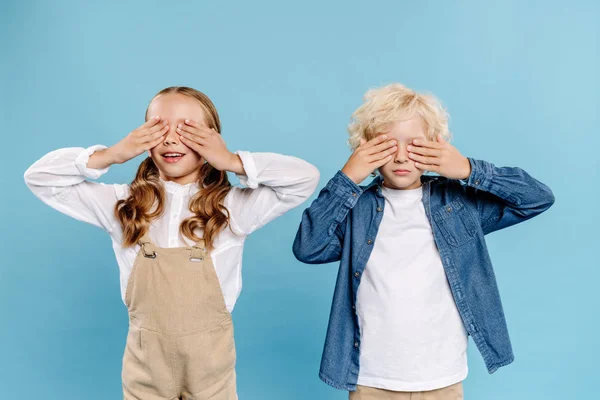 Smiling kids obscuring faces with hands isolated on blue — Stock Photo