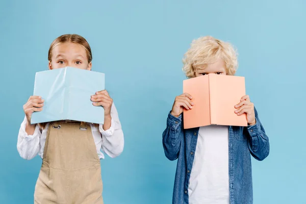 Niños mirando a la cámara y oscureciendo caras con libros aislados en azul - foto de stock