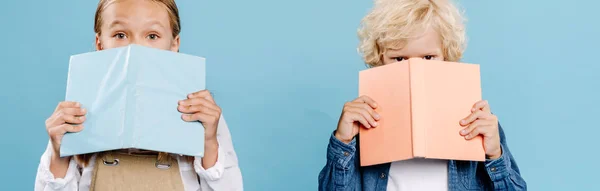 Panoramic shot of kids looking at camera and obscuring faces with books isolated on blue — Stock Photo