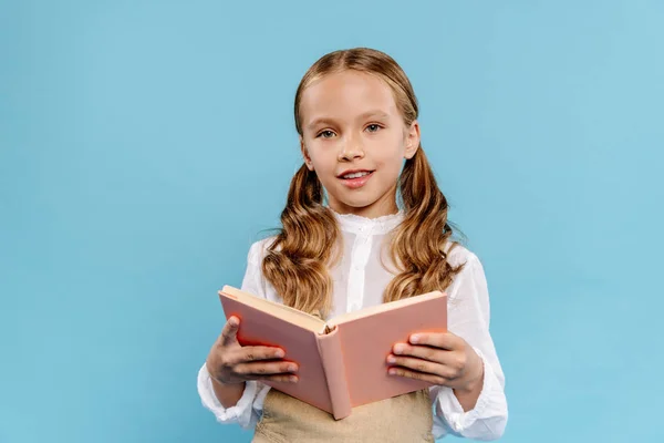 Lächelndes und nettes Kind, das in die Kamera schaut und Buch isoliert auf blauem Grund hält — Stockfoto