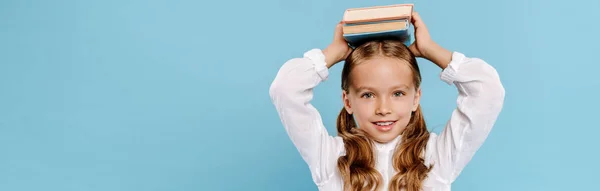Tiro panorâmico de criança sorrindo e bonito olhando para a câmera e segurando livros isolados em azul — Fotografia de Stock