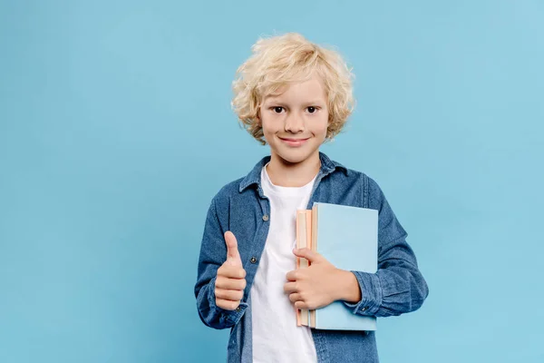 Sonriente y lindo niño mostrando como gesto y sosteniendo libros aislados en azul — Stock Photo