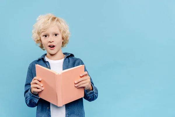 Schockiertes und nettes Kind, das Buch in der Hand hält und isoliert auf blau in die Kamera schaut — Stockfoto