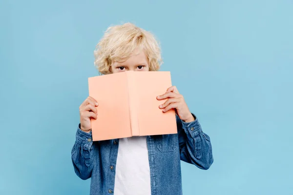Bonito garoto obscurecendo rosto com livro isolado no azul — Fotografia de Stock