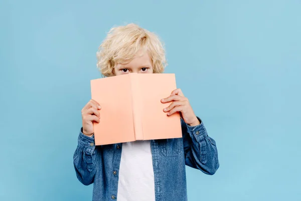 Lindo niño oscureciendo la cara con libro aislado en azul - foto de stock