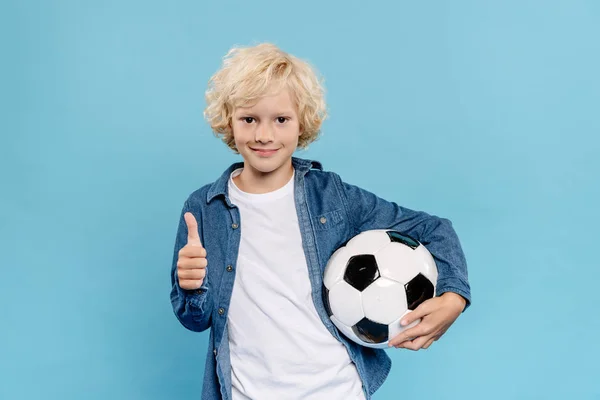 Sorrindo e bonito garoto mostrando como e segurando futebol isolado em azul — Fotografia de Stock