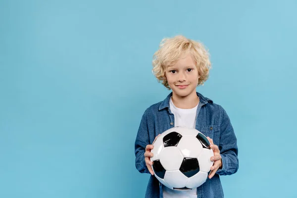 Sonriente y lindo niño mirando a la cámara y sosteniendo el fútbol aislado en azul — Stock Photo