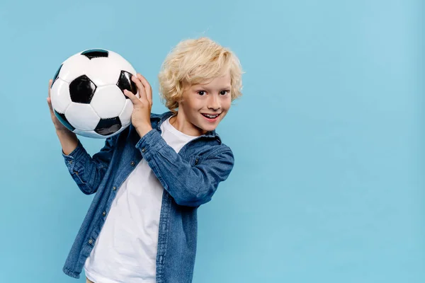 Lächelndes und süßes Kind, das in die Kamera schaut und den Fußball isoliert auf blauem Grund hält — Stockfoto