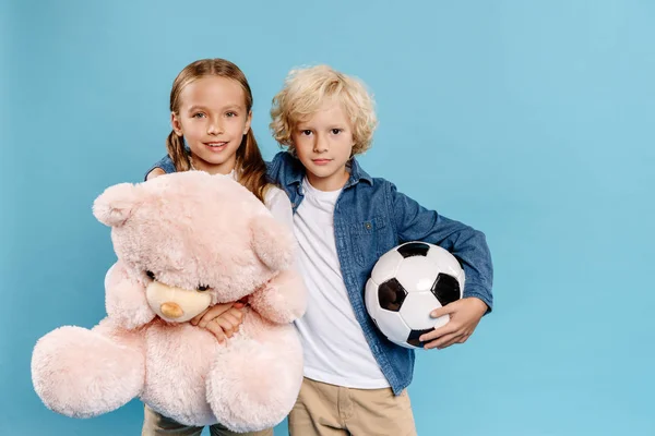 Sorridente e bonito crianças segurando ursinho de pelúcia e futebol isolado em azul — Fotografia de Stock