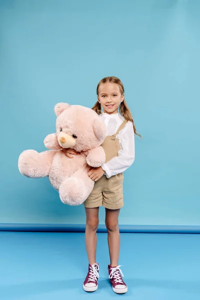 Sonriente y lindo niño mirando a la cámara y sosteniendo oso de peluche sobre fondo azul - foto de stock