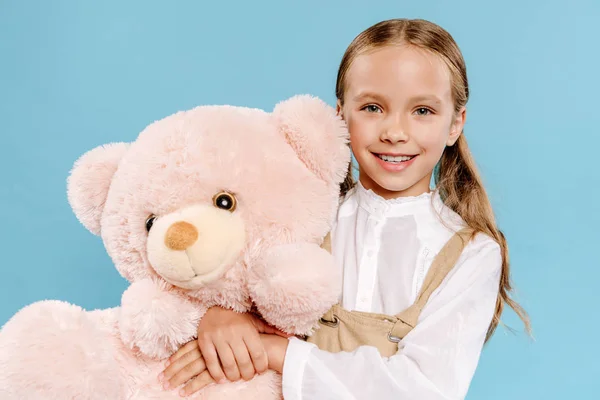 Smiling and cute kid looking at camera and holding teddy bear isolated on blue — Stock Photo