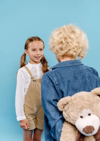 Rückansicht des Jungen mit Teddybär und lächelndem Kind, das ihn isoliert auf blau betrachtet — Stockfoto