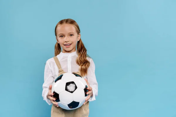 Lächelndes und süßes Kind, das in die Kamera schaut und den Fußball isoliert auf blauem Grund hält — Stockfoto
