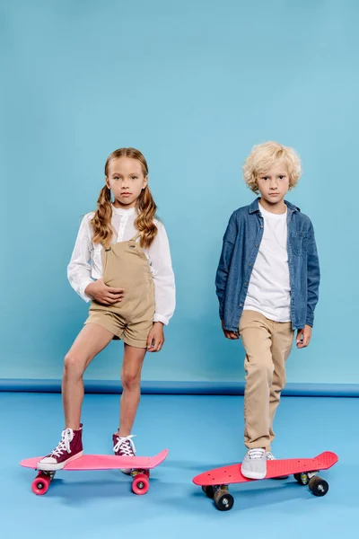 Niedliche Kinder, die in die Kamera schauen und auf Penny-Brettern auf blauem Hintergrund stehen — Stockfoto