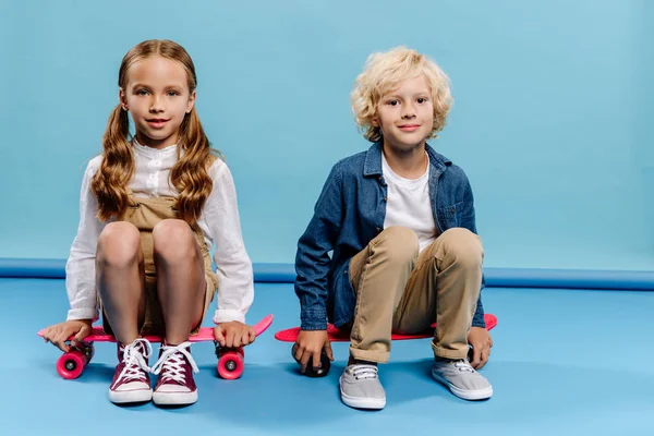 Lächelnde und niedliche Kinder, die in die Kamera schauen und auf Penny-Brettern auf blauem Hintergrund sitzen — Stockfoto